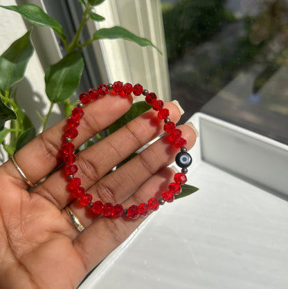 A red beaded bracelet with a black evil eye charm, held in a person's hand near a window with green leaves in the background.