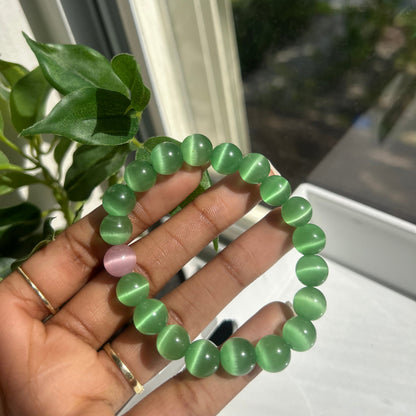 A beaded bracelet made of green cat's eye stones with a single pink accent bead, held in a person's hand near a window.