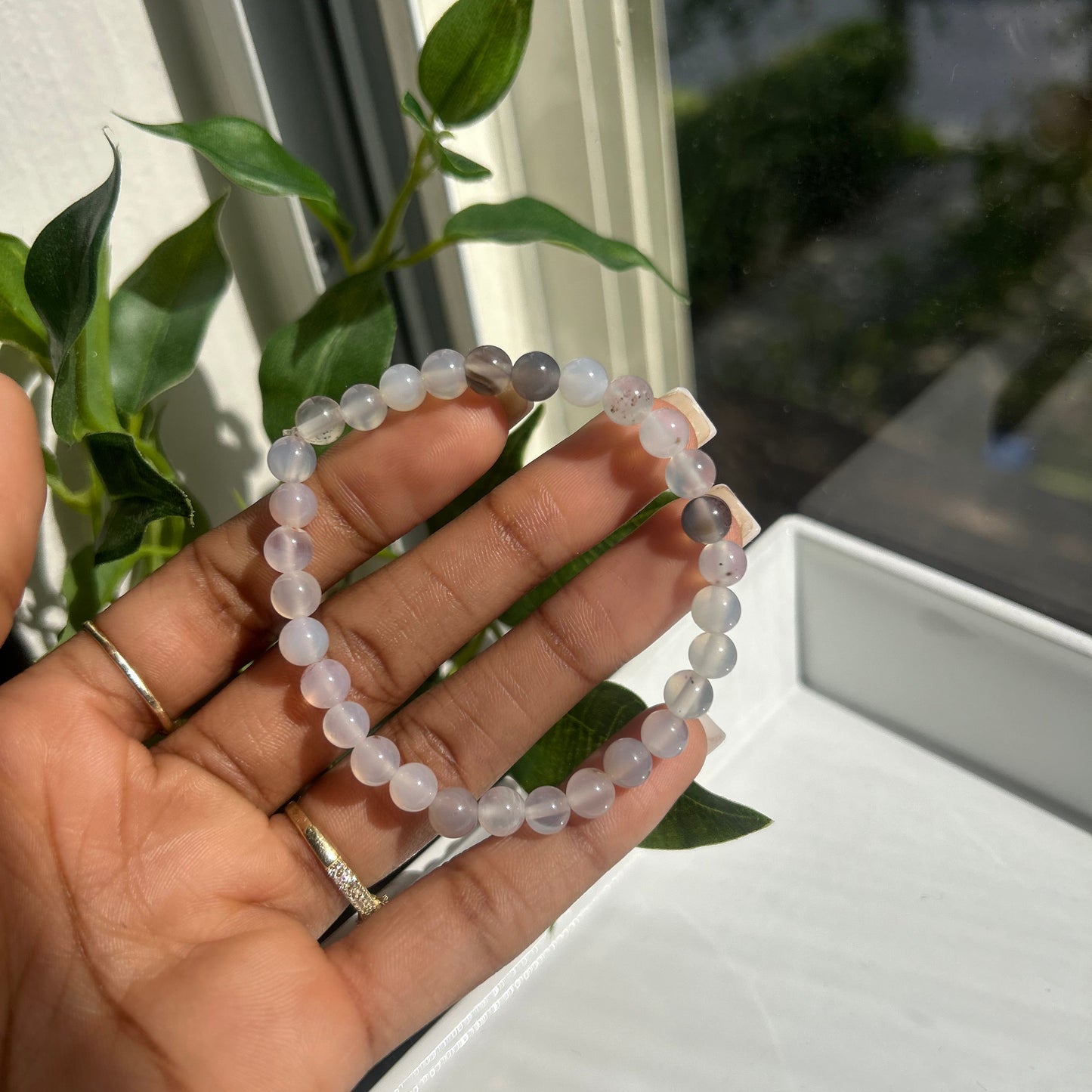 A beaded bracelet made of pale lavender-gray translucent round beads, held in a person's hand near a window with green leaves in the background.