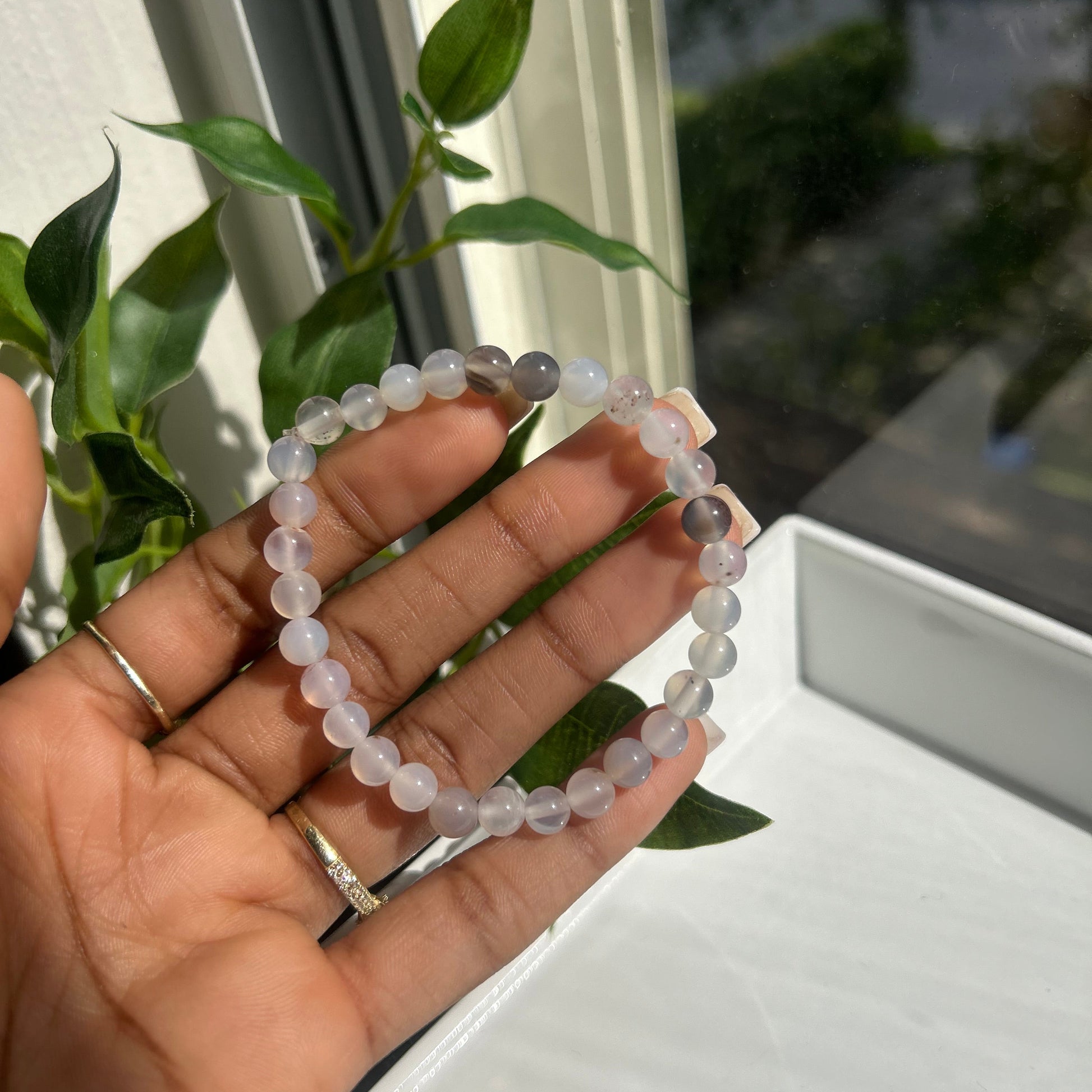 A beaded bracelet made of pale lavender-gray translucent round beads, held in a person's hand near a window with green leaves in the background.