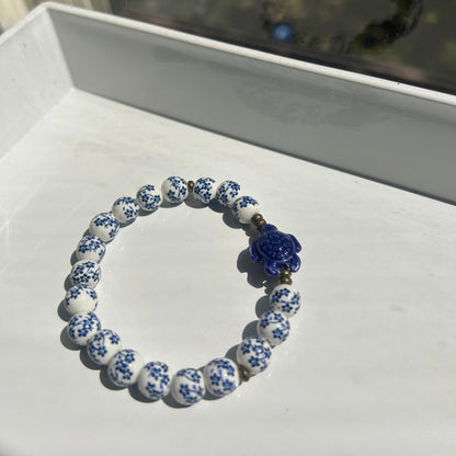 A beaded bracelet with white and blue patterned round beads and a central blue flower charm, displayed on a white surface.