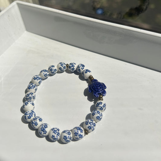 A beaded bracelet with white and blue patterned round beads and a central blue flower charm, displayed on a white surface.