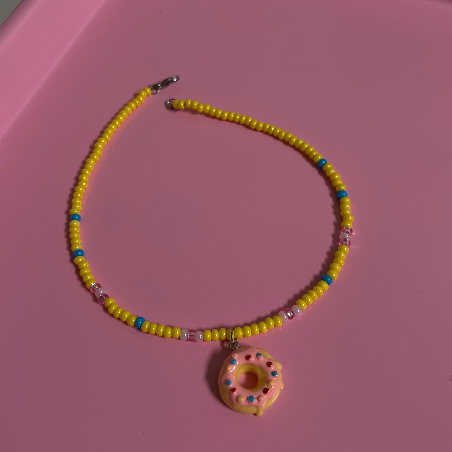 Yellow beaded necklace with a pink donut charm on a pink surface