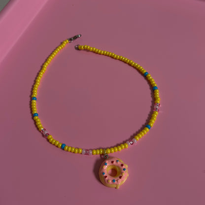 Yellow beaded necklace with a pink donut charm on a pink surface
