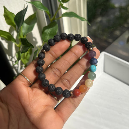 A beaded stretch bracelet held in a hand, featuring dark gray lava stone beads on one side and a rainbow of colored gemstone beads on the other, with a small silver charm in the center.